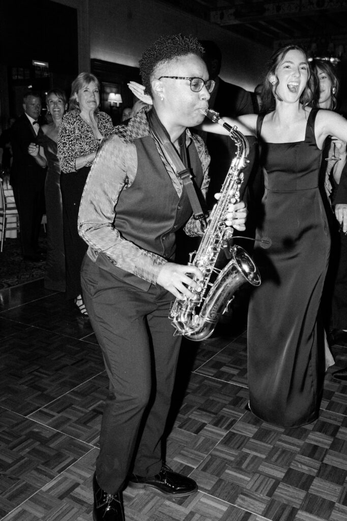 Live saxophone musician performing on the dance floor while wedding guests cheer and dance around him during a lively Detroit wedding reception. The energetic black-and-white image captures the excitement and celebration that comes with getting married in Detroit, highlighting the vibrant party atmosphere couples look for when choosing where to get married in Detroit MI. Reception entertainment photographed by Detroit local photographer Courtney Rudicel.