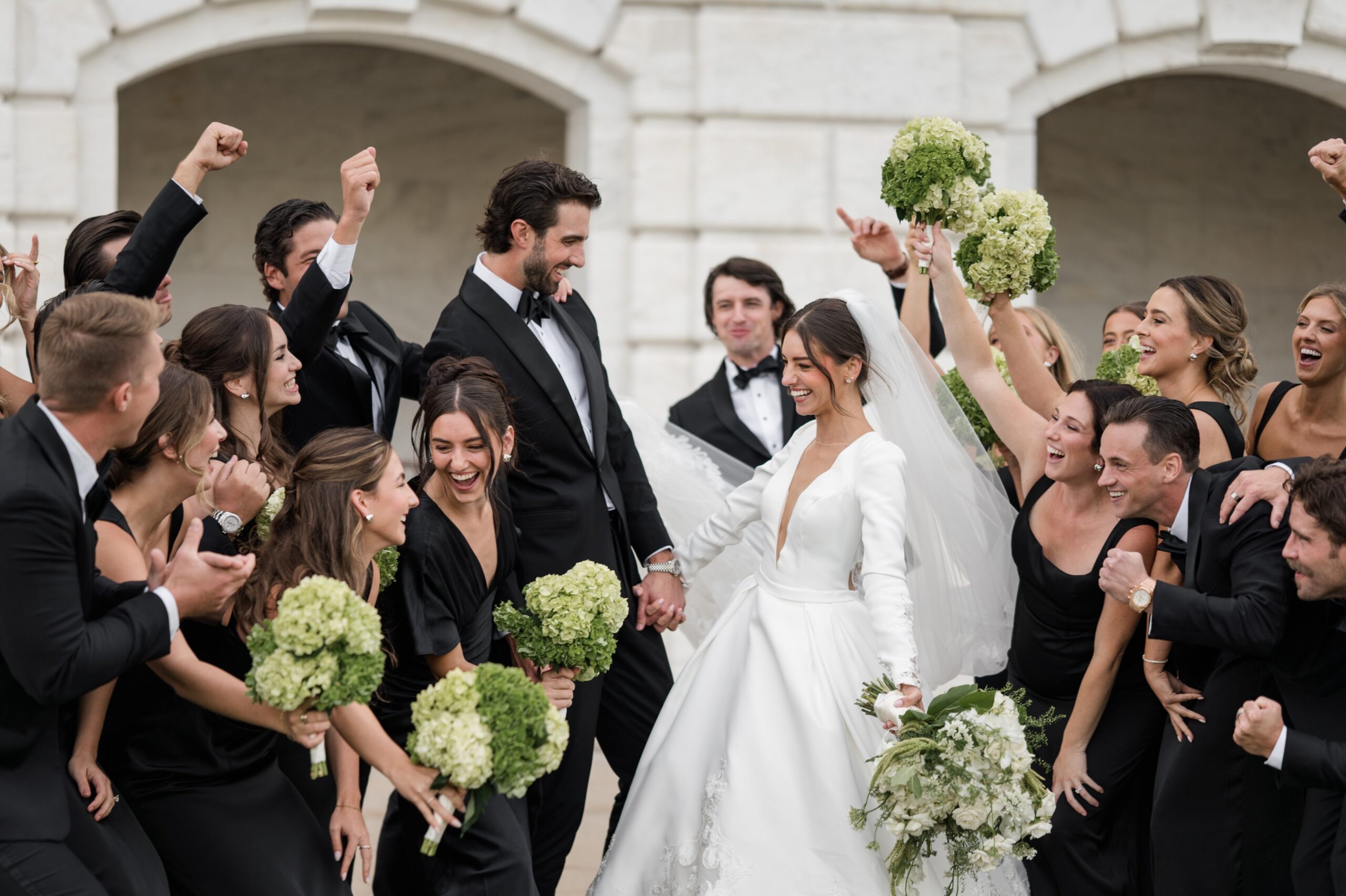 Bride and groom celebrating with their wedding party outside a historic Detroit building, surrounded by bridesmaids in black dresses holding green bouquets and groomsmen in black tuxedos cheering with raised arms. The bride smiles while holding a white floral bouquet and wearing a long-sleeved gown and veil, capturing the joyful energy of getting married in Detroit Michigan at one of the beautiful places to get married in Detroit MI. Candid Detroit wedding moment photographed by local photographer Courtney Rudicel, showing laughter, movement, and celebration among close friends and family while getting married in Detroit.