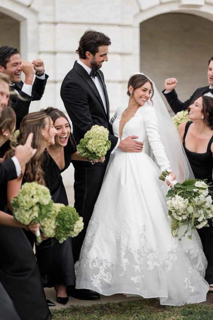 Bride and groom celebrate outdoors with their wedding party, smiling and laughing while bridesmaids hold green hydrangea bouquets around the couple. Joyful group portrait capturing the excitement of getting married in Detroit during a stylish Detroit wedding photographed by local photographer Courtney Rudicel.