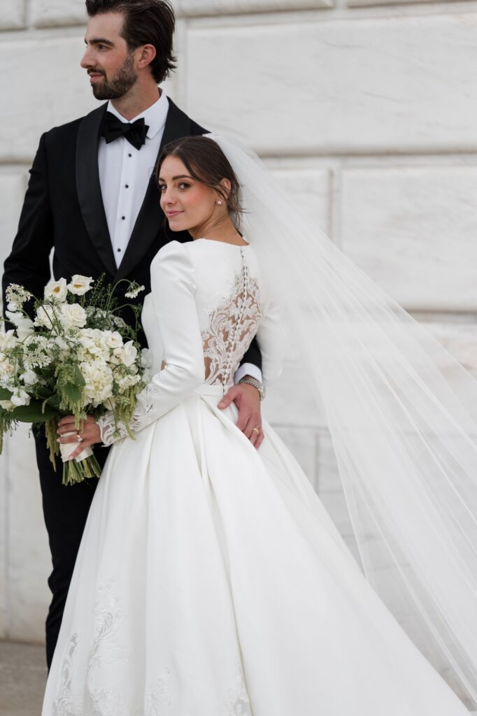Bride and groom posing together against a marble architectural backdrop, the bride holding a white floral bouquet while her cathedral veil flows behind her and the groom stands in a classic black tuxedo. The romantic portrait reflects the sophistication of a Detroit wedding and showcases the beautiful city architecture couples seek when deciding where to get married in Detroit. Wedding portrait photographed by Detroit local photographer Courtney Rudicel.