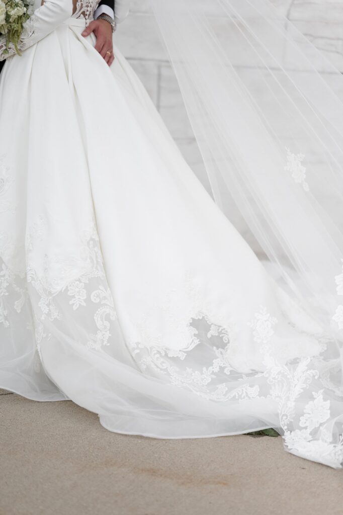 Close-up detail of a bride’s flowing wedding gown and cathedral-length veil draped across the ground, highlighting delicate lace embroidery and timeless bridal fashion during a Detroit wedding portrait session. Elegant bridal detail photographed while getting married in Detroit Michigan by Courtney Rudicel.