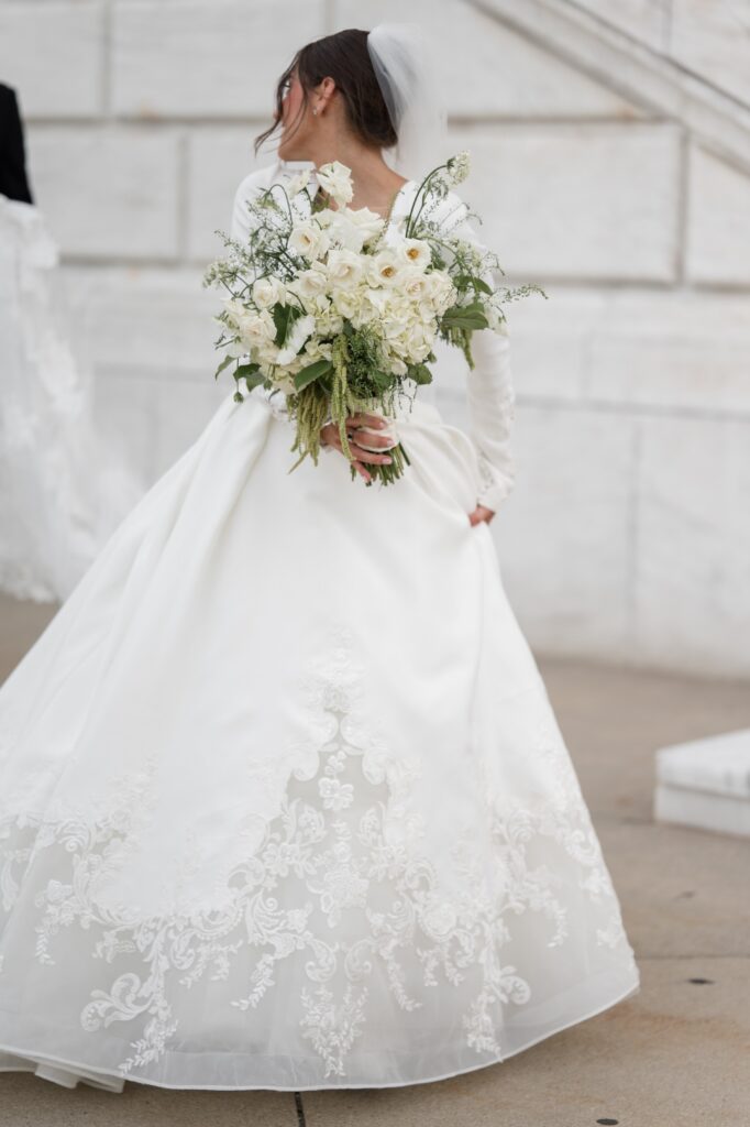 Bride standing outdoors holding a lush white bouquet behind her back, showcasing the full silhouette and lace detailing of her wedding dress against classic stone architecture. Romantic bridal portrait representing where to get married in Detroit MI, photographed by Detroit wedding photographer Courtney Rudicel.