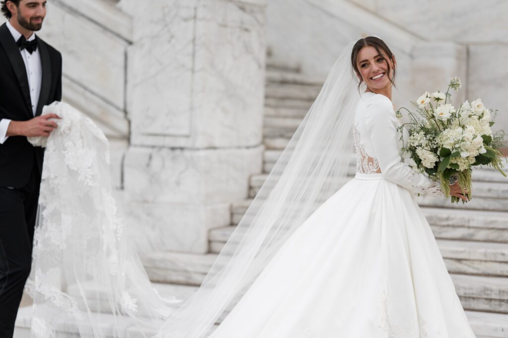 Bride smiling over her shoulder while holding a white floral bouquet as the groom gently lifts her cathedral-length veil on marble steps outside a historic Detroit venue. The romantic portrait highlights the elegance of getting married in Detroit Michigan, showcasing beautiful architectural backdrops couples seek when choosing where to get married in Detroit MI. Detroit wedding portrait photographed by local photographer Courtney Rudicel.