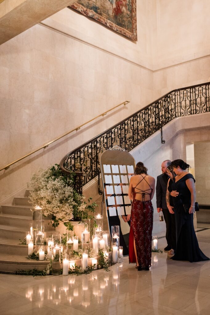 Wedding guests reviewing a seating chart display beside a grand marble staircase decorated with greenery arrangements and glowing candles inside an elegant Detroit venue. The sophisticated reception entrance highlights thoughtful wedding design details often seen at luxury places to get married in Detroit MI, capturing the welcoming atmosphere of getting married in Detroit. Detroit wedding reception photograph by local photographer Courtney Rudicel.