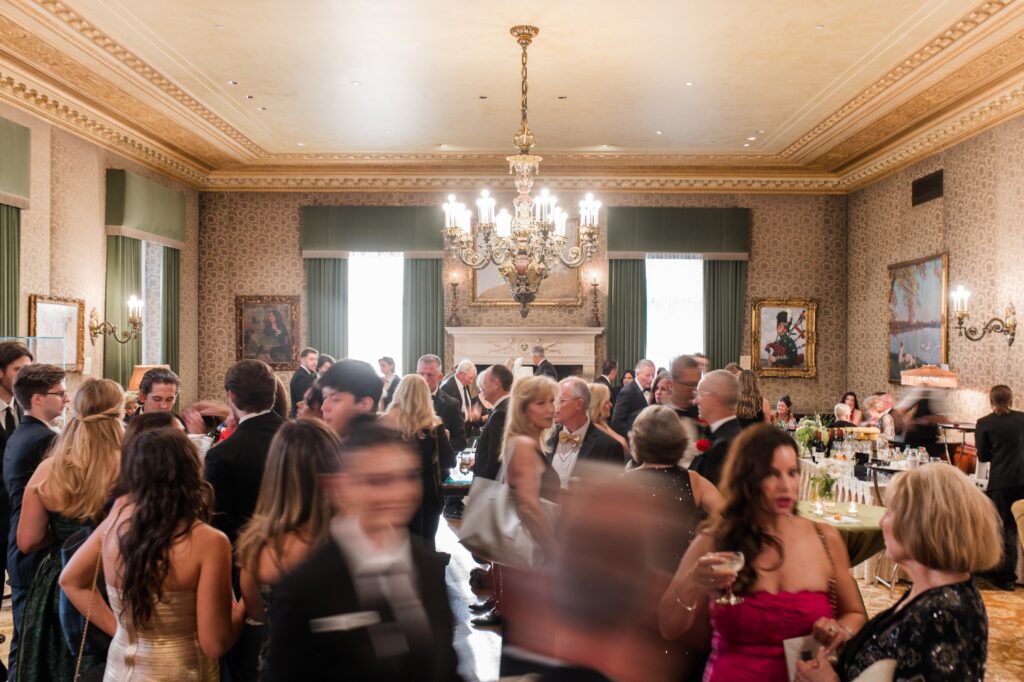 Guests mingling and enjoying cocktails inside an elegant Detroit wedding reception room featuring chandeliers, framed artwork, and classic historic décor. The lively cocktail hour scene captures friends and family socializing before dinner, reflecting the sophisticated experience of getting married in Detroit Michigan and showcasing one of the refined places to get married in Detroit MI. Detroit wedding reception moment photographed by local photographer Courtney Rudicel.