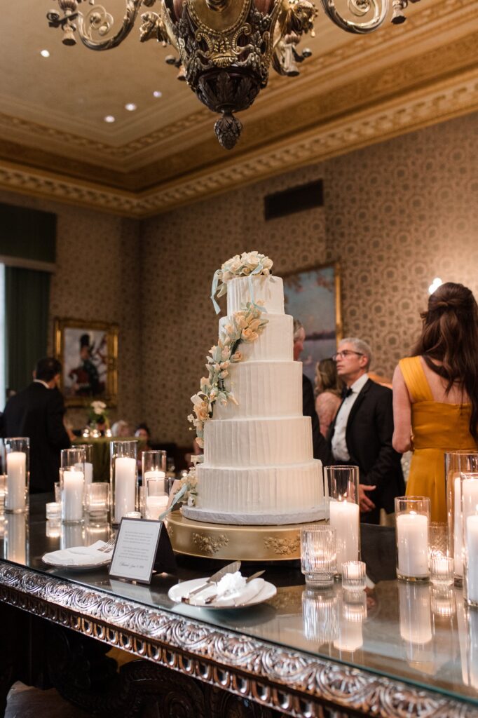 Four-tier white wedding cake decorated with delicate sugar flowers and displayed on a mirrored table surrounded by candlelight inside a historic Detroit reception space. Guests mingle in the background beneath ornate chandeliers, showcasing refined reception styling typical of a classic Detroit wedding and inspiring couples researching where to get married in Detroit Michigan. Wedding cake detail photographed by Detroit local photographer Courtney Rudicel.