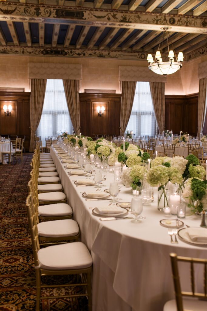 Long reception tables set with white linens, gold chairs, candlelight, and green hydrangea centerpieces inside an elegant wood-paneled Detroit ballroom with decorative ceiling beams and chandeliers. The formal dining setup illustrates one of the timeless options for where to get married in Detroit MI, highlighting sophisticated reception design for couples getting married in Detroit. Detroit wedding reception setup photographed by local photographer Courtney Rudicel.