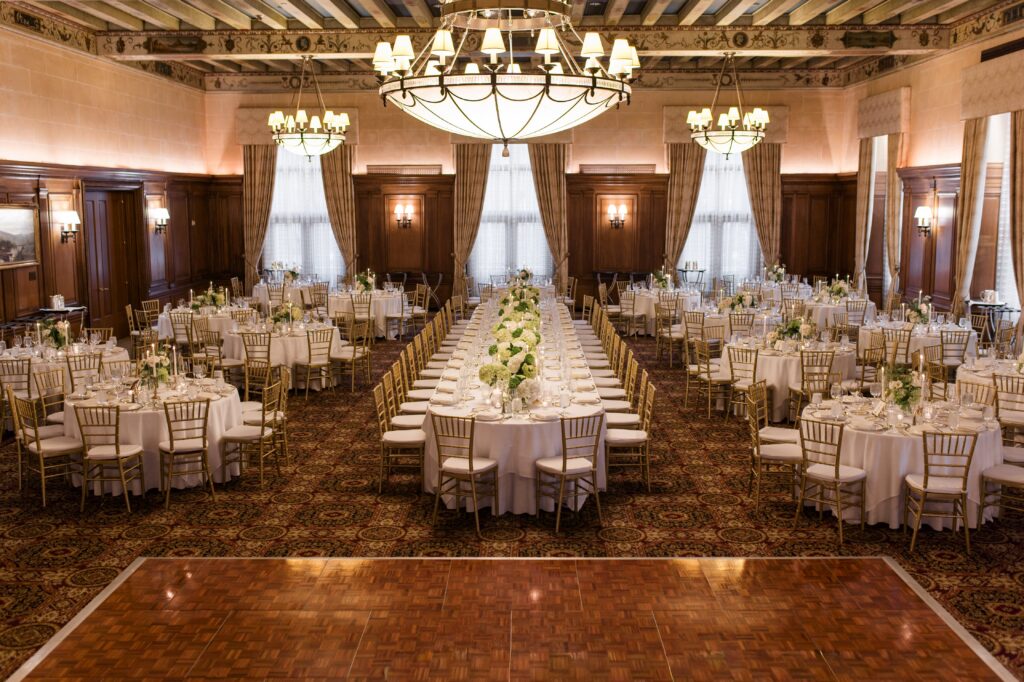 Wide view of an elegant Detroit wedding reception ballroom featuring round guest tables, long banquet tables with green hydrangea centerpieces, gold Chiavari chairs, chandeliers, and a polished dance floor ready for celebration. The refined reception layout showcases one of the timeless places to get married in Detroit MI, helping couples envision getting married in Detroit Michigan in a classic and sophisticated venue setting. Detroit wedding reception space photographed by local photographer Courtney Rudicel.