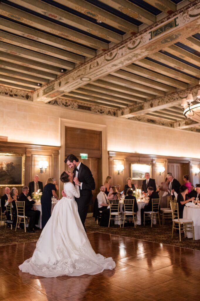 Bride and groom sharing their first dance on a polished ballroom floor surrounded by seated wedding guests inside an elegant Detroit reception venue with wood paneling and decorative ceiling beams. The romantic moment captures the joy of a classic Detroit wedding and reflects the timeless atmosphere couples imagine when getting married in Detroit Michigan. First dance photograph by local Detroit wedding photographer Courtney Rudicel.