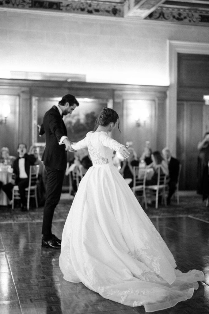 Bride and groom dancing together on a polished ballroom floor during their first dance, the bride’s full gown flowing as guests watch from surrounding tables. The elegant black-and-white moment reflects the romance and tradition of a classic Detroit wedding, inspiring couples planning getting married in Detroit Michigan at timeless reception venues. First dance photograph captured by local Detroit wedding photographer Courtney Rudicel.