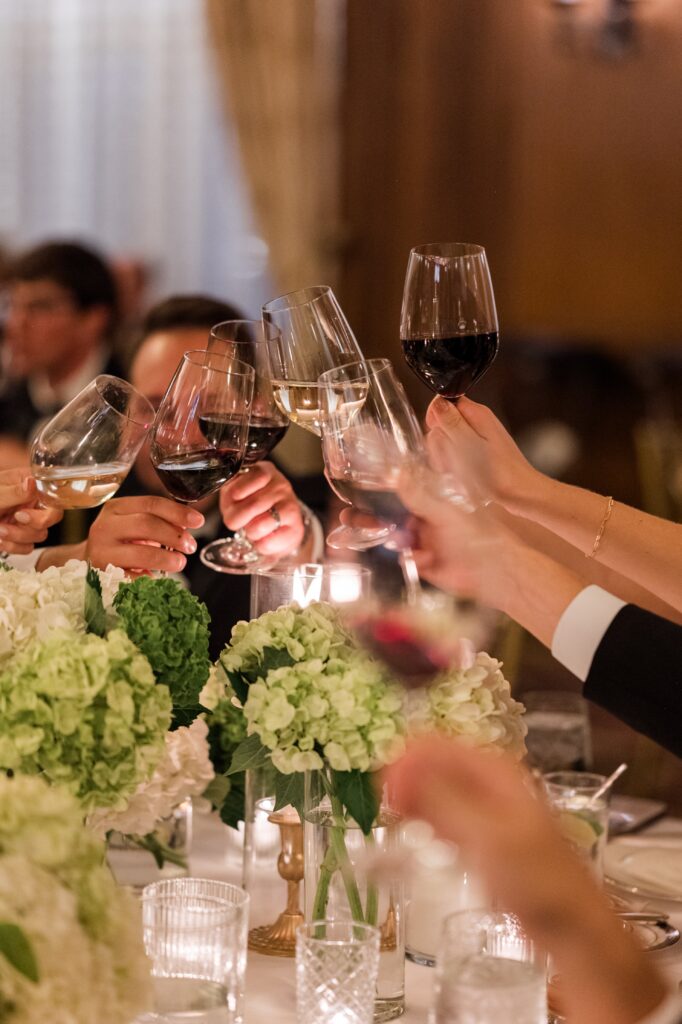Wedding guests raising glasses of red and white wine in a celebratory toast around a reception table decorated with green hydrangea centerpieces and candlelight inside an elegant Detroit venue. The joyful reception moment captures friends and family celebrating together during a classic Detroit wedding, reflecting the lively atmosphere couples experience when getting married in Detroit Michigan. Reception photograph by local Detroit wedding photographer Courtney Rudicel.