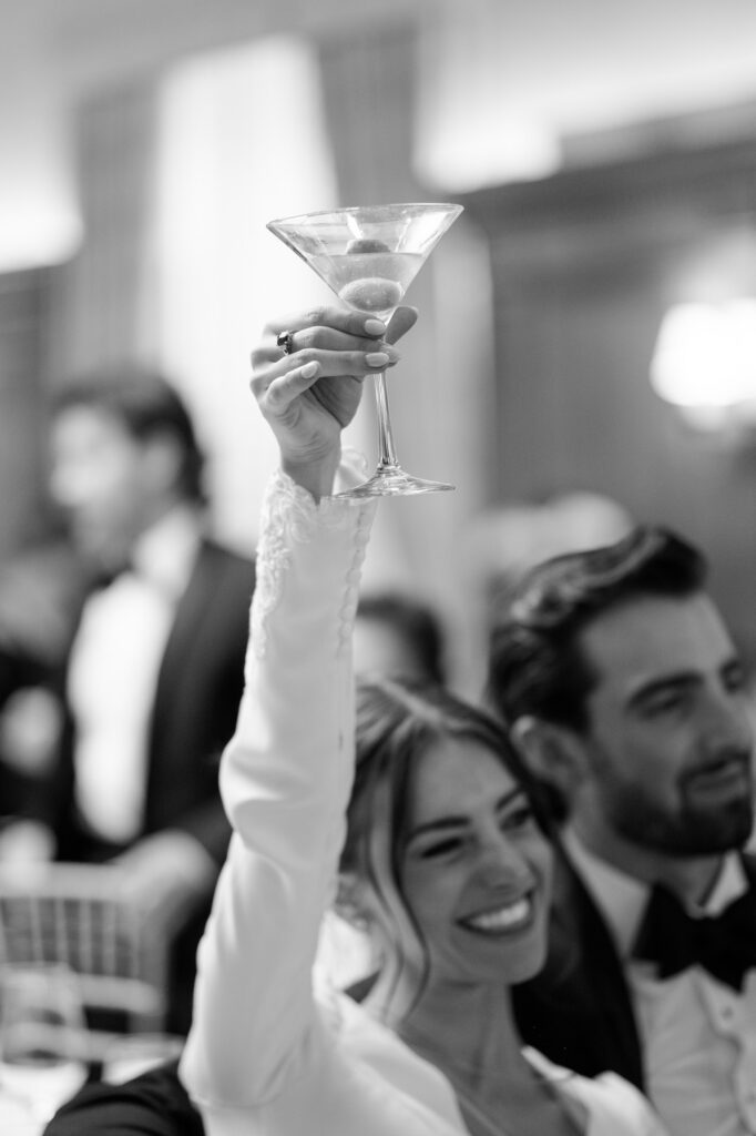 Bride smiling and lifting a martini glass for a toast during the wedding reception, surrounded by softly blurred guests dressed in formal attire. The candid black-and-white image highlights the celebration and emotion of getting married in Detroit, showcasing the festive energy found at many places to get married in Detroit MI. Documentary-style Detroit wedding photograph by local photographer Courtney Rudicel.
