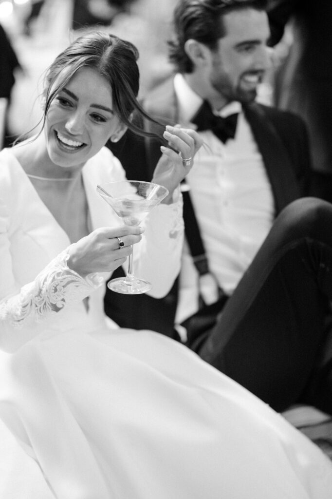 Bride laughing while holding a martini glass and sitting beside the groom during the reception, captured in an intimate black-and-white moment filled with joy and relaxation after the ceremony. The candid interaction reflects the fun and personal side of a Detroit wedding, inspiring couples researching where to get married in Detroit MI for an elegant yet lively celebration. Wedding reception image photographed by Detroit local photographer Courtney Rudicel.