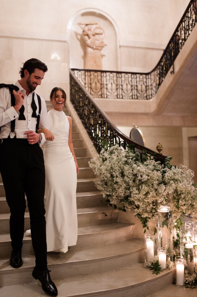 Bride and groom walking down a grand marble staircase decorated with white florals and glowing candles, smiling together during their wedding reception entrance. The sophisticated venue design highlights one of the luxurious places to get married in Detroit MI, showcasing the elegance couples seek when planning where to get married in Detroit. Detroit wedding reception photograph by local photographer Courtney Rudicel.