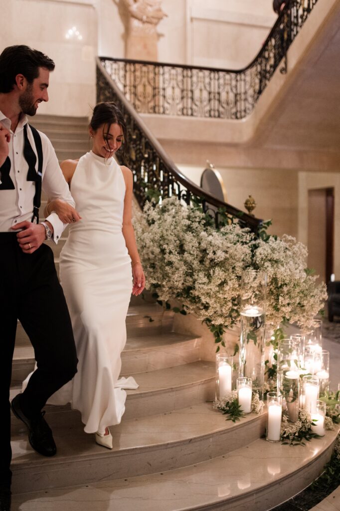 Newly married couple descending a candlelit staircase hand in hand, surrounded by romantic floral arrangements and historic architectural details inside a Detroit wedding venue. The intimate moment reflects the celebration and excitement of getting married in Detroit, illustrating an elegant reception experience for couples researching where to get married in Detroit MI. Wedding reception image photographed by Detroit local photographer Courtney Rudicel.