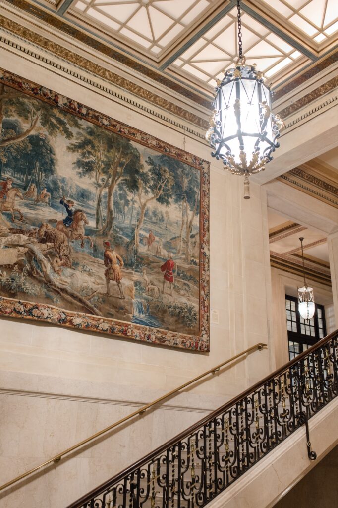 Grand staircase inside a historic Detroit wedding venue with wrought iron railing, large hanging lantern chandelier, and a wall-sized tapestry depicting a pastoral scene. The refined architectural atmosphere demonstrates where to get married in Detroit MI for couples seeking elegant indoor ceremony and reception spaces while getting married in Detroit Michigan. Detroit wedding venue image photographed by local photographer Courtney Rudicel.