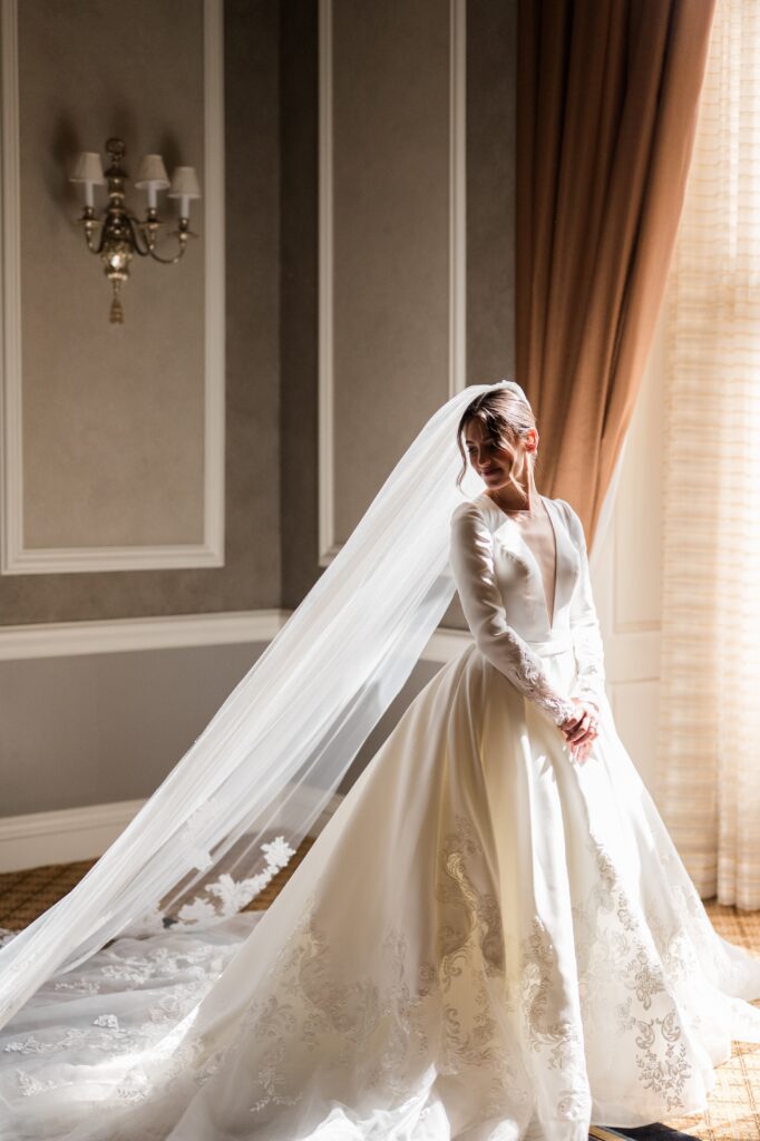 Bride standing near a sunlit window wearing a long-sleeved white wedding gown with a flowing cathedral veil trailing across the floor, softly illuminated by natural light inside a classic Detroit venue. The quiet portrait captures a peaceful bridal moment before the ceremony, reflecting the elegance of a Detroit wedding and inspiring couples researching where to get married in Detroit. Bridal portrait photographed by Detroit local photographer Courtney Rudicel.