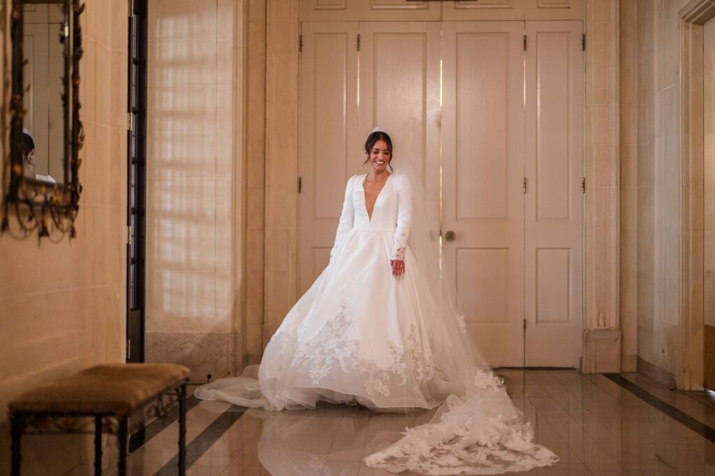 Bride standing in a grand historic Detroit hallway wearing a long-sleeved wedding gown with lace detailing and a flowing cathedral-length veil, smiling softly as her dress moves around her. The elegant bridal portrait highlights the timeless beauty of getting married in Detroit and showcases one of the sophisticated indoor settings couples consider when deciding where to get married in Detroit MI. Detroit wedding portrait photographed by local photographer Courtney Rudicel.