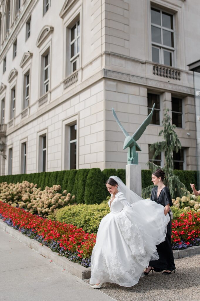 Bride walking outside a historic Detroit wedding venue while a bridesmaid helps carry her flowing veil, surrounded by manicured gardens and classic architecture. The candid outdoor moment highlights the elegance of getting married in Detroit Michigan and showcases beautiful exterior spaces couples consider when choosing where to get married in Detroit. Outdoor Detroit wedding photograph captured by local photographer Courtney Rudicel.