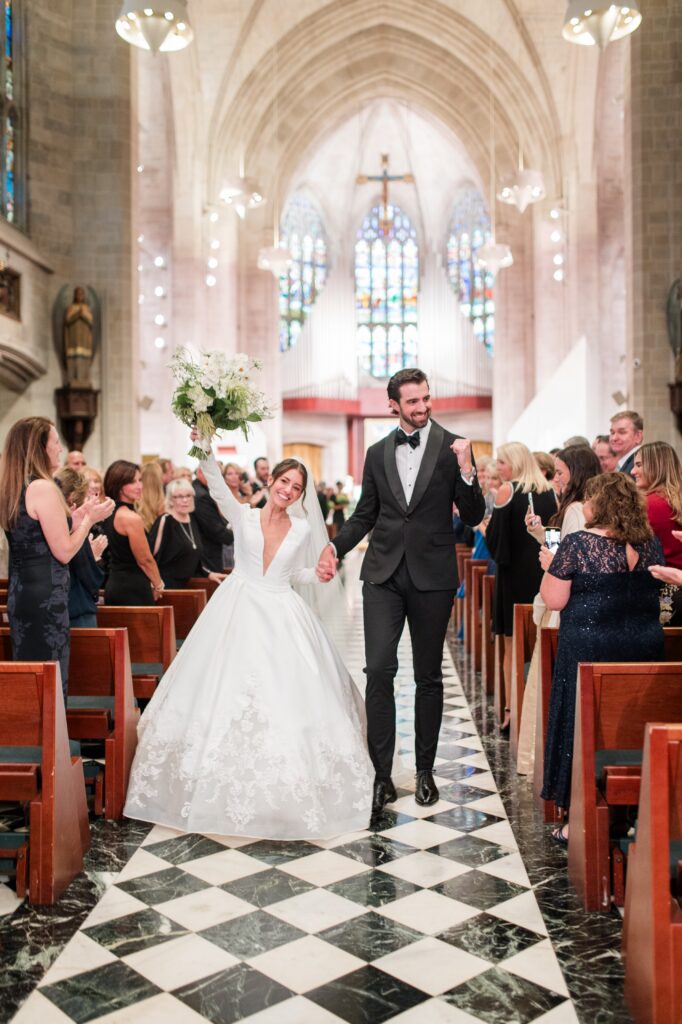 Newly married couple walking joyfully down the church aisle after their ceremony, the bride raising her bouquet while guests stand and applaud from wooden pews beneath stained glass windows. The celebratory recessional captures the excitement of getting married in Detroit and reflects the emotional moment couples experience when deciding where to get married in Detroit MI for a meaningful ceremony. Detroit wedding ceremony photographed by local photographer Courtney Rudicel.