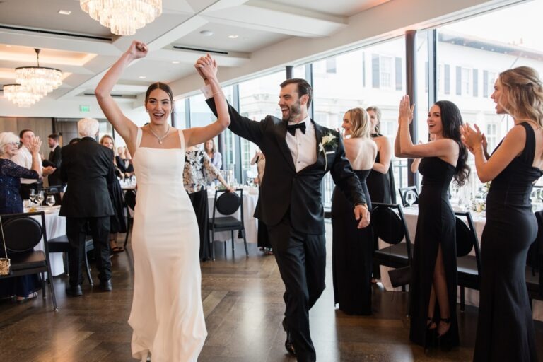 Bride and groom smiling and raising their hands as they enter their wedding reception surrounded by cheering guests
