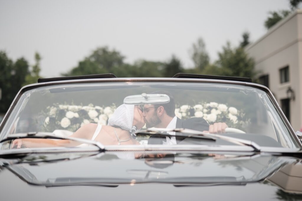 Bride and groom kissing in the front seat of a vintage black convertible during their wedding portraits.
