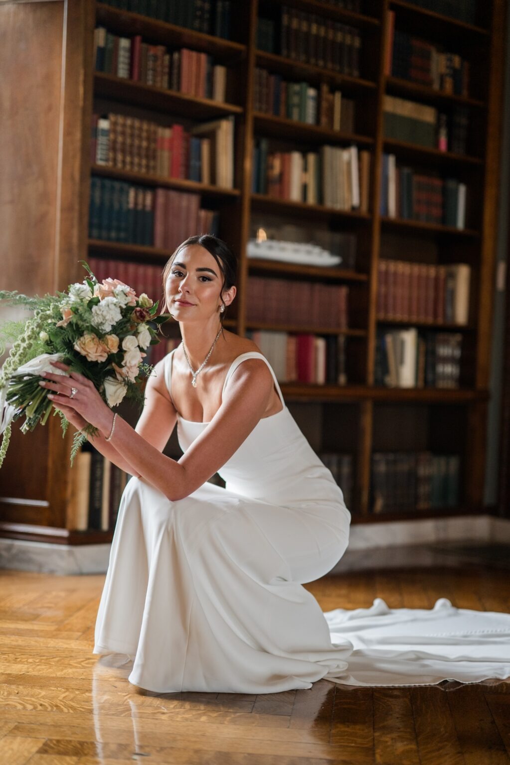 Bride holding her peach and white wedding bouquet in front of the bookshelves inside the Alger House library
