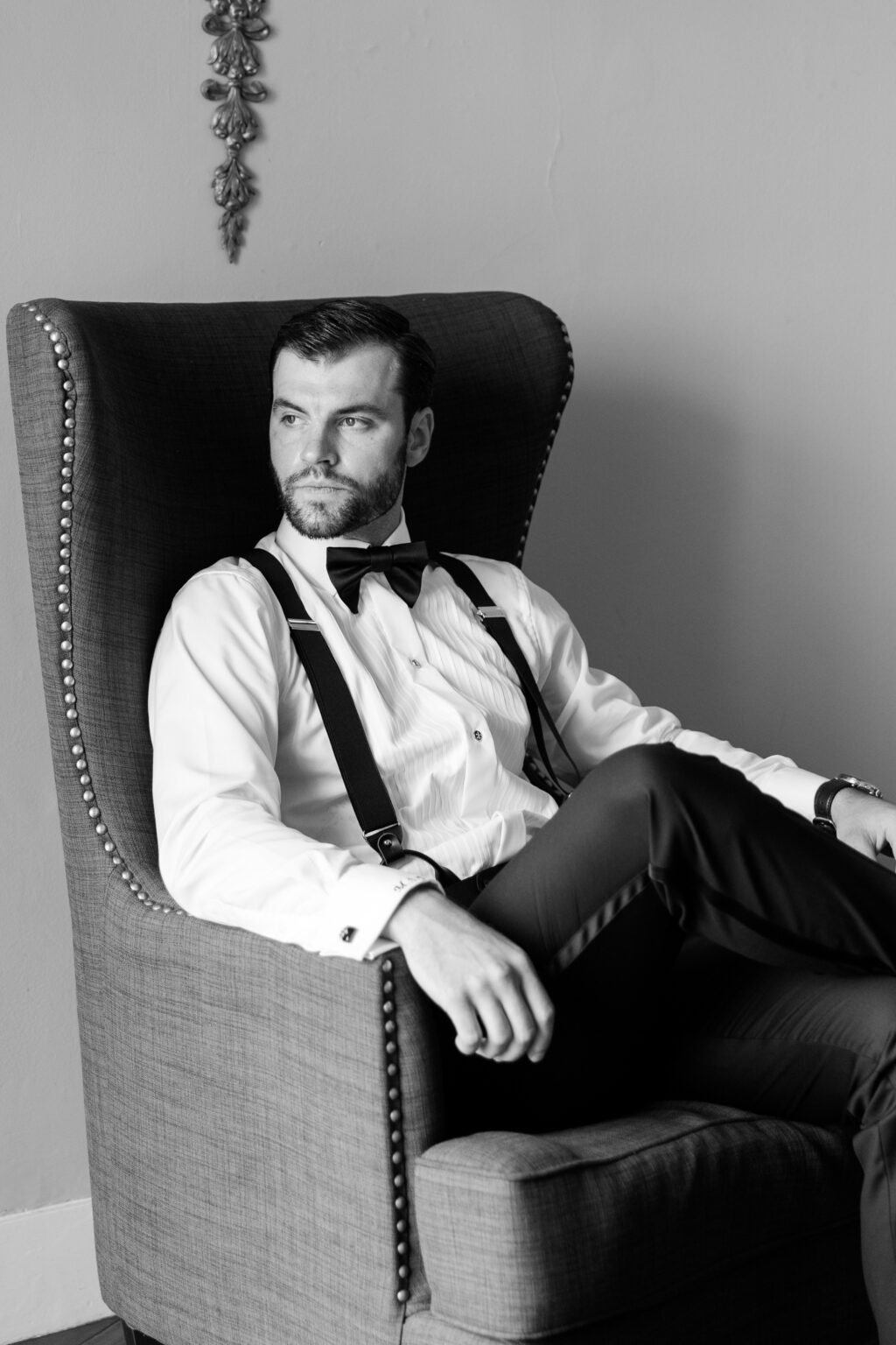 Black and white portrait of Maxx seated in a wingback chair wearing suspenders and a black bow tie before the wedding